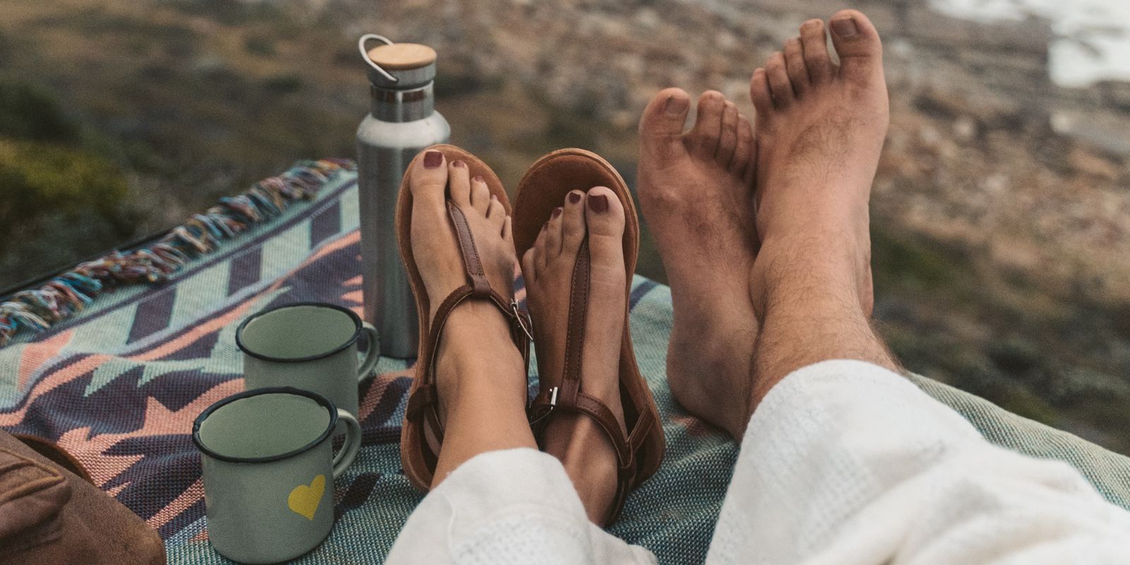 Female legs with sandals right next to a man who is barefeet and has them crossed over each other sitted in a picnic setting